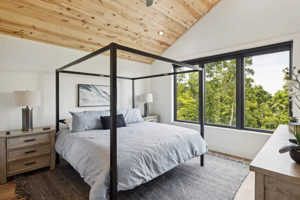 A serene bedroom featuring a four-poster bed placed in front of large windows that frame lush greenery outside. The room is accented by a wooden ceiling and nightstands, creating a tranquil, cozy retreat that feels both spacious and private.
