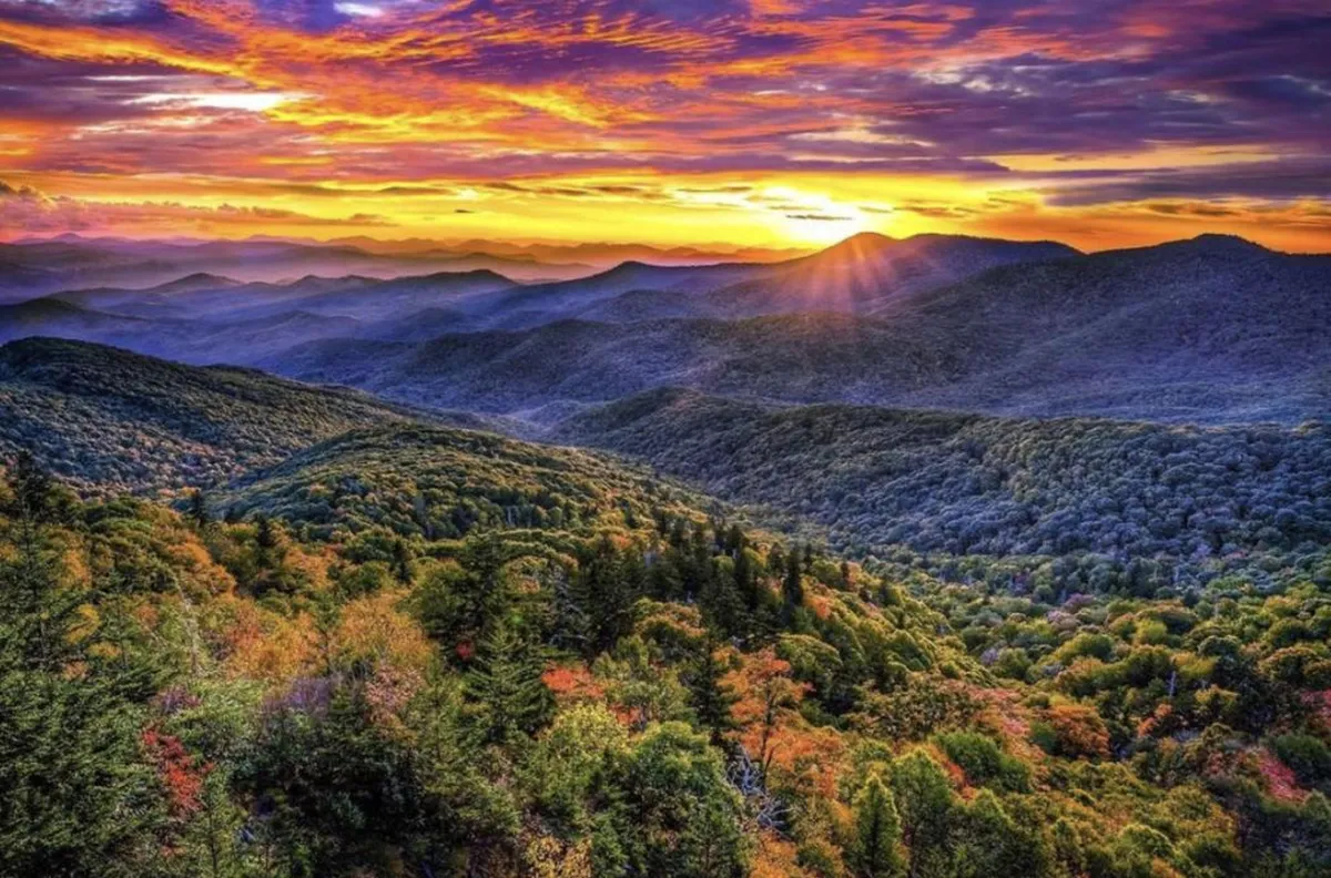 A sweeping mountain landscape at sunset with colorful skies and dense forests, inviting feelings of tranquility and awe. The view captures the natural beauty surrounding Mountain Breeze and evokes the quiet, sophisticated atmosphere promised by the property.