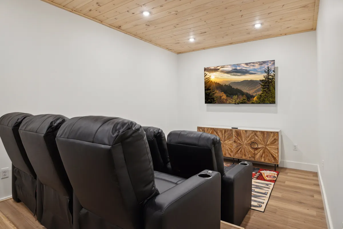 A home theater setup with plush reclining chairs arranged in rows facing a large TV displaying a scenic landscape. Warm wood cabinetry and soft lighting create a cozy, immersive environment for movie nights or game-day gatherings.