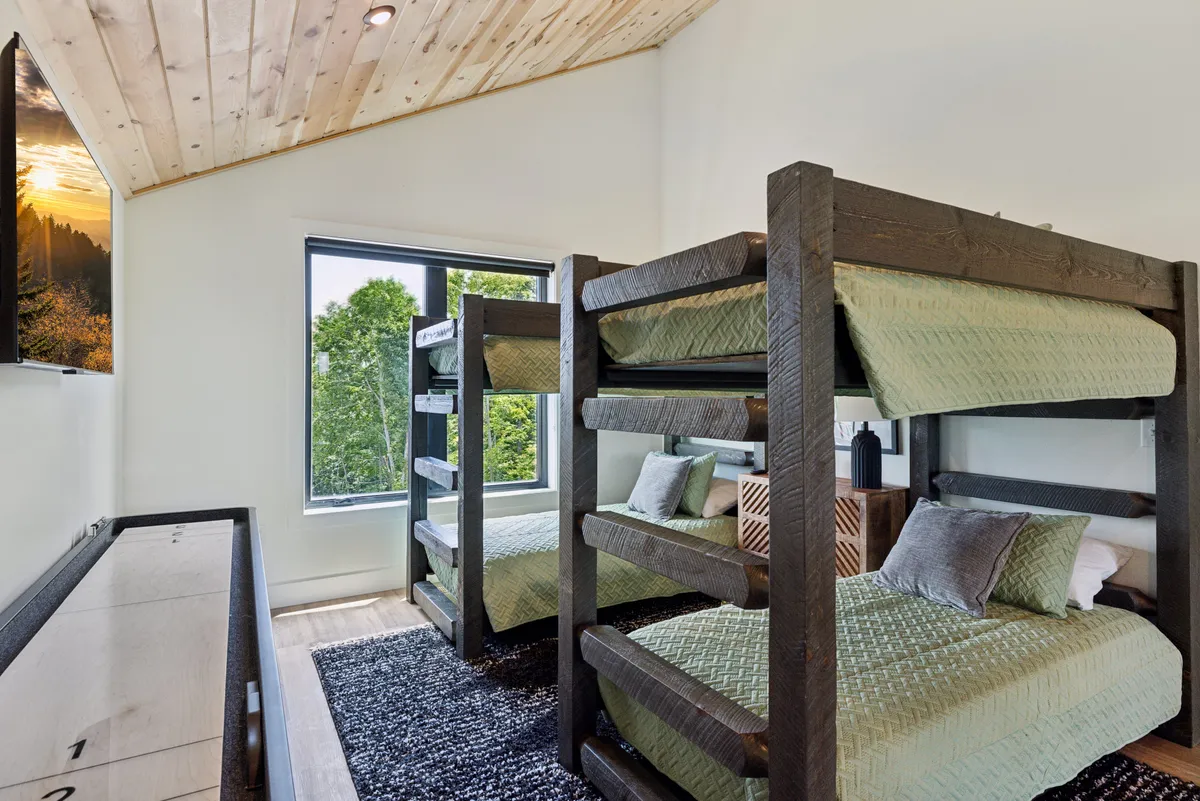 A playful bunk room with twin bunk beds, a large window letting in natural light, a wooden ceiling, and a wall-mounted television. The space is designed for both sleep and leisure, ideal for kids or groups of friends.