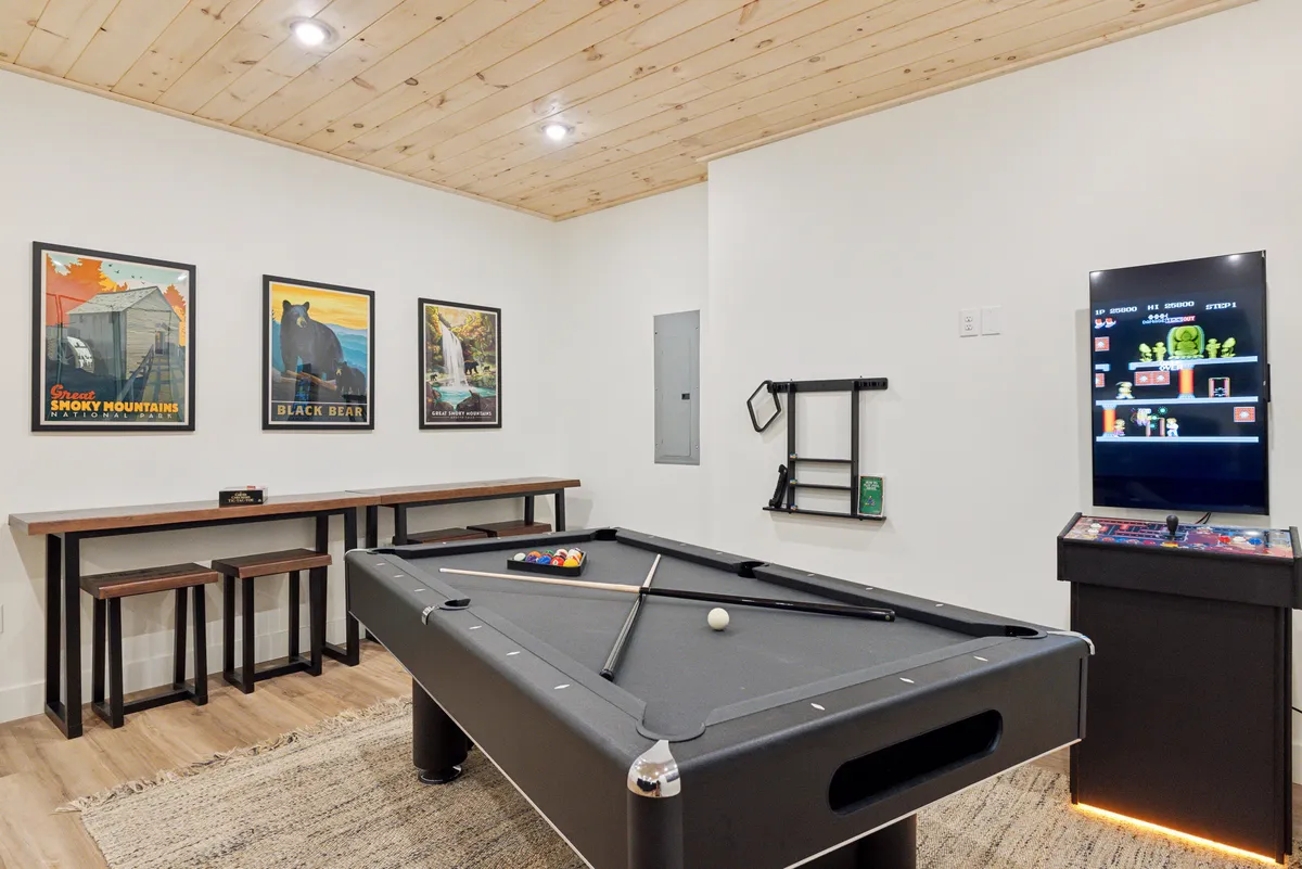 A spacious game room with a classic pool table at its center, complemented by a modern bar table, a video game console, and vibrant wall art. The space is well-lit and designed for both competitive play and relaxed socializing.