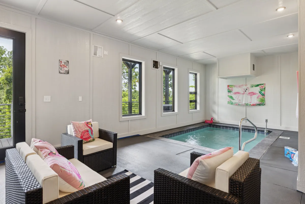 A stylish indoor pool area featuring a sleek pool, black and white striped rug, comfortable seating, and colorful artwork. Large windows let in natural light, ensuring the space feels both private and inviting.