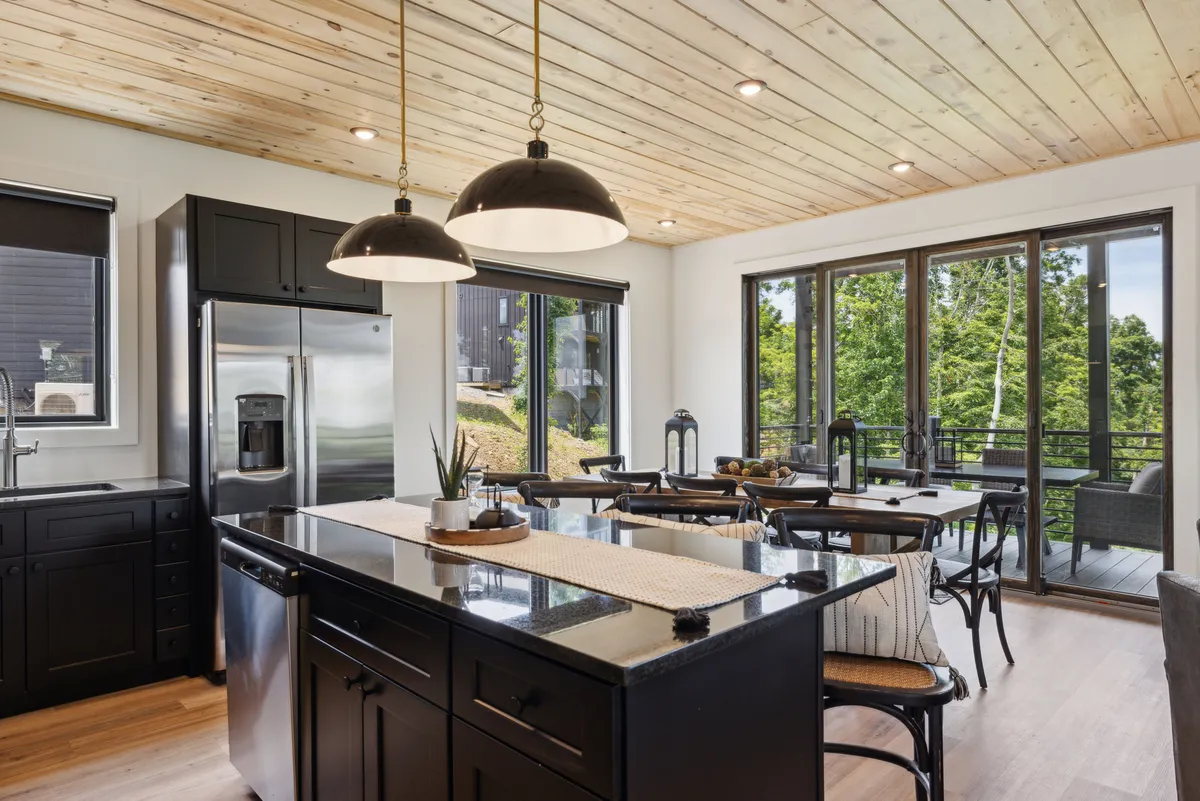 A contemporary kitchen setup with a striking black kitchen island, sleek pendant lighting above, expansive windows inviting natural light, and a glimpse of the adjacent dining area. Modern appliances and ample counter space are visible, emphasizing the gourmet kitchen experience.
