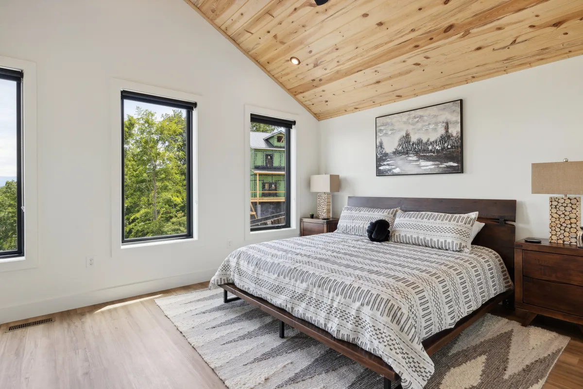 A bright, tranquil bedroom with a comfortable bed, artwork above the headboard, windows letting in natural light, a cozy rug, and a bedside lamp. The room’s soft colors and modern décor promote a sense of calm and relaxation.