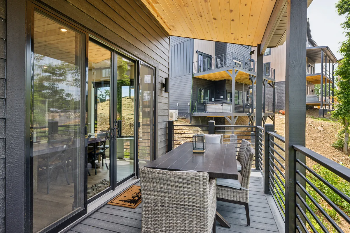 This exterior image highlights an inviting deck with an outdoor dining table, wicker chairs, a wooden ceiling, and sliding glass doors. It's arranged for social gatherings, meals, and entertaining, all with a backdrop of natural beauty. The setting is cozy and encourages group interactions in the fresh air.