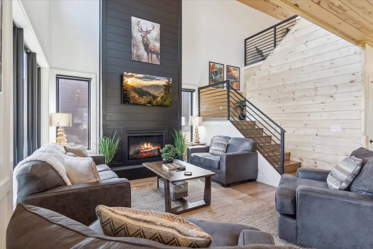 This image features a cozy living room with a prominent fireplace, large windows that frame scenic views, a modern staircase, artwork on the walls, and a central coffee table. Sunlight streams in, creating a warm, inviting space ideal for relaxation and socializing.